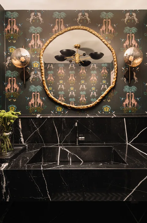 A Mumbai apartment powder room with black marble, patterned wallpaper, a round brass mirror and decorative lighting