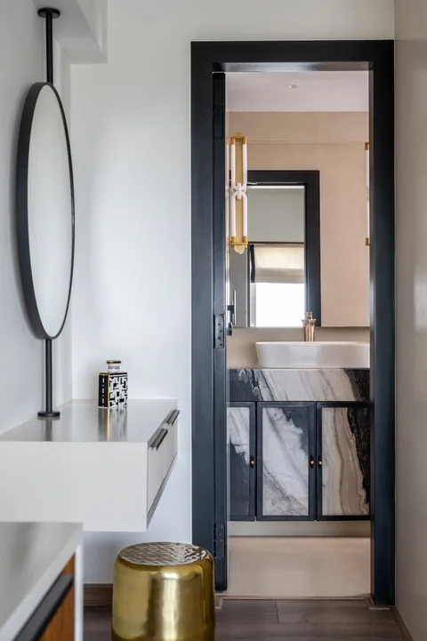 A Mumbai apartment bathroom with a slim white vanity, round mirror, brass bin and a view through the doorway